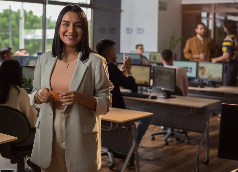 Mujer sonriendo en una oficina, con compañeros de trabajo al fondo.