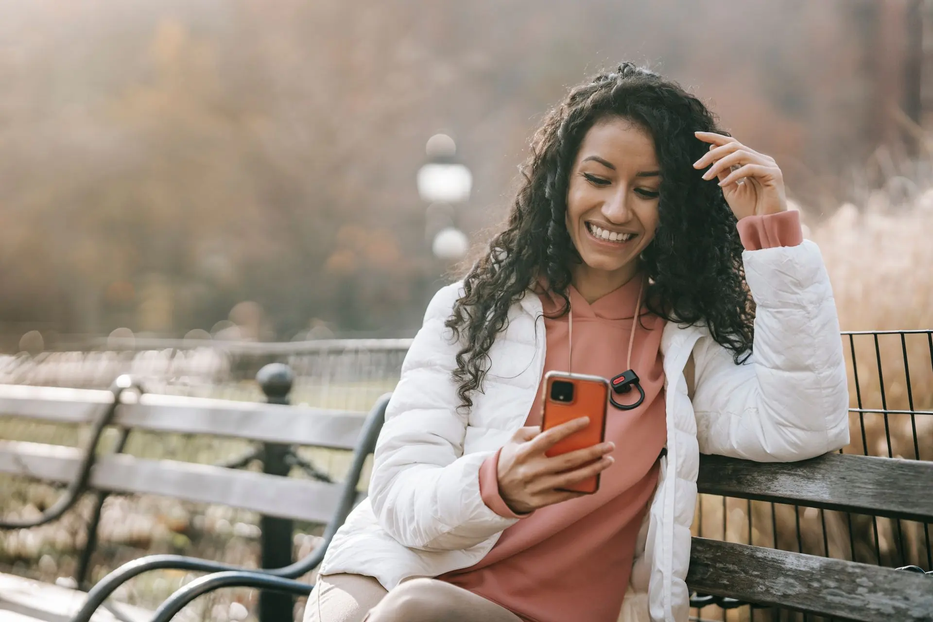Mujer sonriendo mientras revisa su teléfono, sentada en una banca al aire libre.